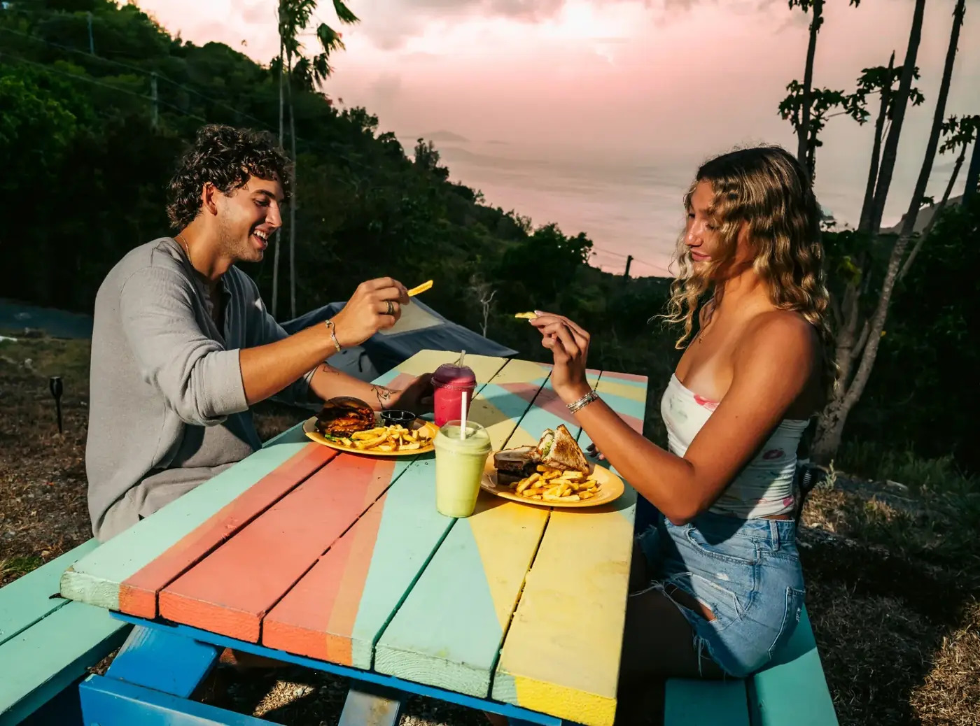 Guests enjoying burgers and smoothies at a colorful Drifters picnic table at sunset on St. John